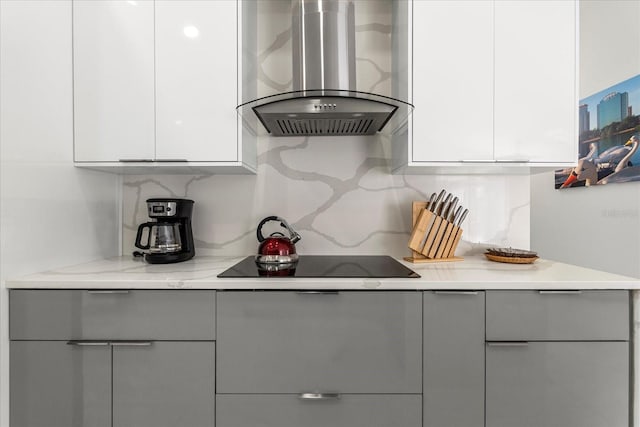 kitchen with light stone counters, white cabinets, wall chimney range hood, black electric cooktop, and decorative backsplash