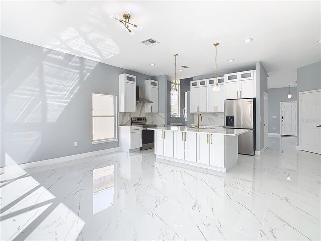 kitchen with wall chimney range hood, white cabinets, hanging light fixtures, a center island with sink, and stainless steel appliances