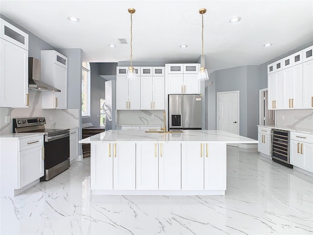 kitchen featuring wall chimney exhaust hood, an island with sink, hanging light fixtures, stainless steel appliances, and beverage cooler