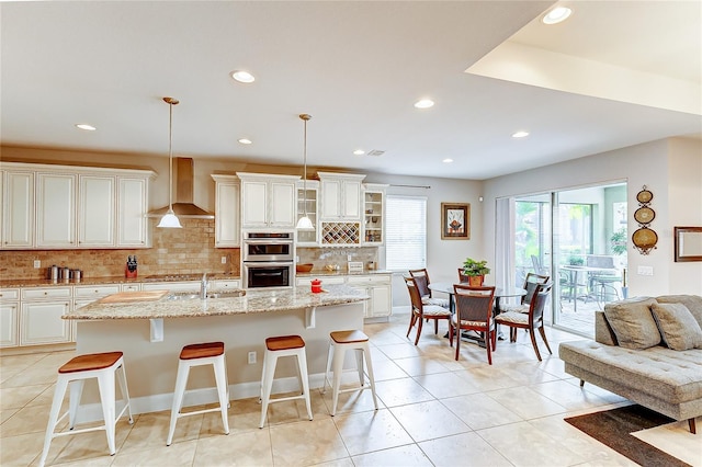 kitchen featuring pendant lighting, wall chimney range hood, backsplash, and an island with sink