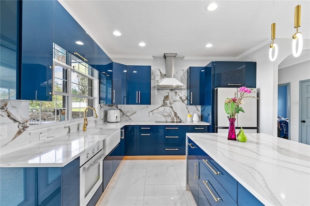 kitchen featuring stainless steel oven, blue cabinets, wall chimney exhaust hood, white fridge, and decorative light fixtures