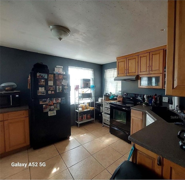 kitchen with dark countertops, under cabinet range hood, a sink, light tile patterned floors, and black appliances