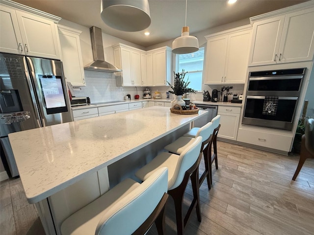 kitchen featuring light stone counters, decorative light fixtures, wall chimney range hood, appliances with stainless steel finishes, and light hardwood / wood-style floors