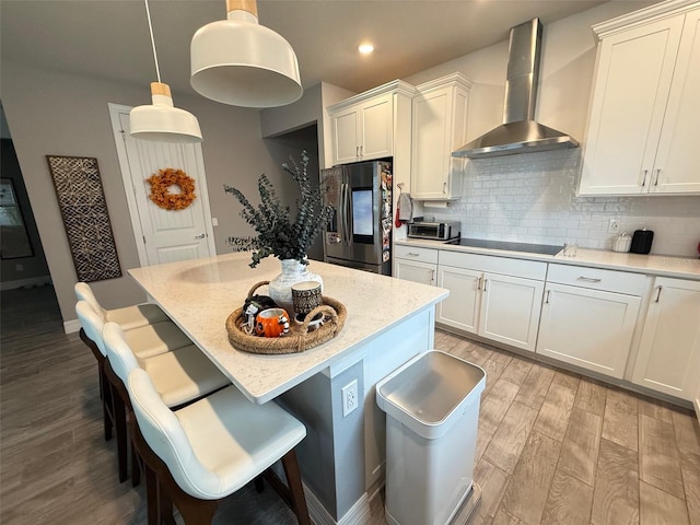 kitchen featuring wall chimney exhaust hood, light wood-type flooring, hanging light fixtures, stainless steel refrigerator with ice dispenser, and black electric stovetop