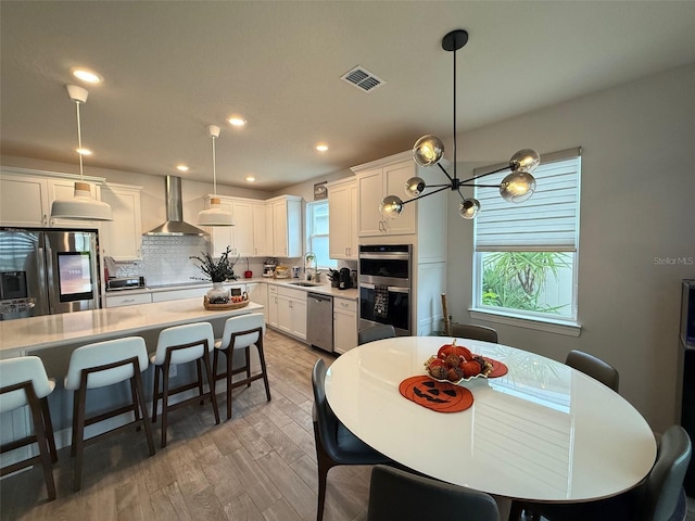 kitchen with wall chimney range hood, pendant lighting, sink, stainless steel appliances, and white cabinetry