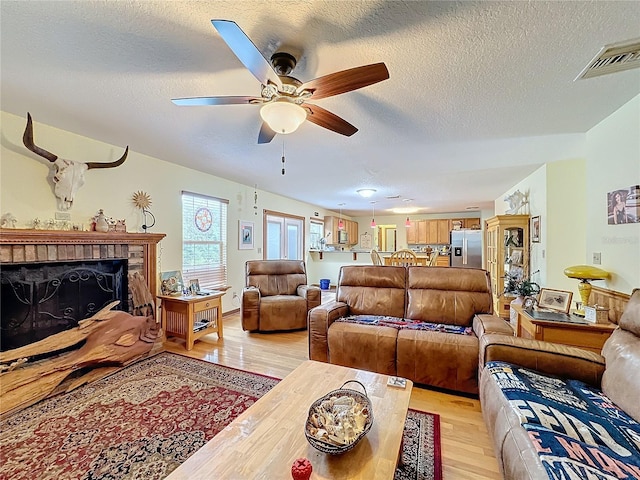 living room with ceiling fan, a brick fireplace, light hardwood / wood-style floors, and a textured ceiling