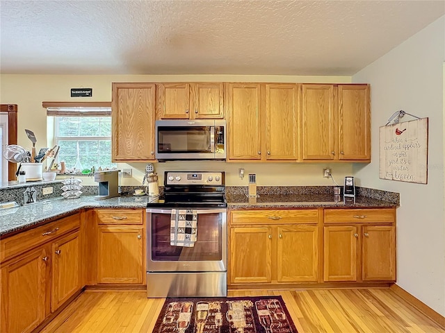 kitchen with a textured ceiling, stainless steel appliances, dark stone counters, and light hardwood / wood-style flooring