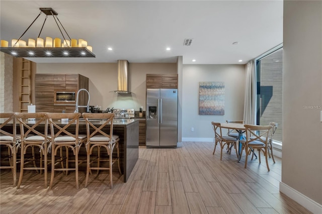 kitchen featuring wall chimney range hood, stainless steel fridge, hanging light fixtures, a kitchen bar, and light hardwood / wood-style flooring