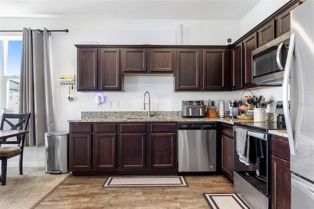 kitchen with dark brown cabinets, light stone countertops, wood-type flooring, sink, and stainless steel appliances