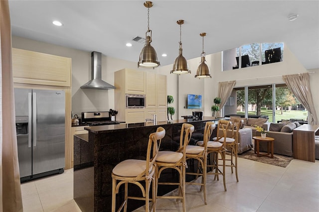 kitchen with appliances with stainless steel finishes, an island with sink, wall chimney exhaust hood, decorative light fixtures, and light tile patterned floors