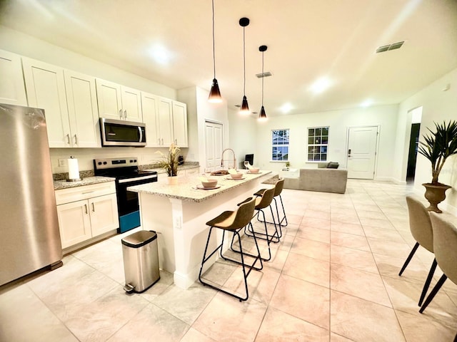 kitchen with pendant lighting, a kitchen island with sink, stainless steel appliances, light stone countertops, and white cabinets