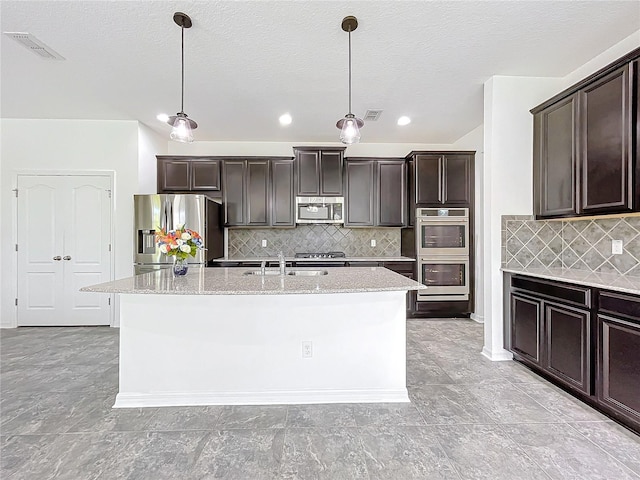 kitchen with decorative backsplash, appliances with stainless steel finishes, dark brown cabinets, a center island with sink, and decorative light fixtures