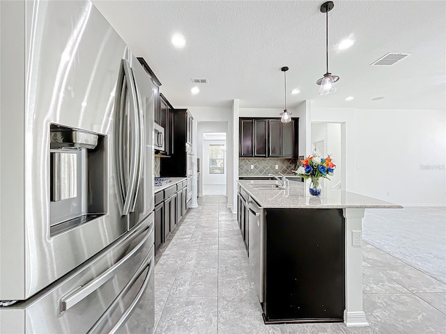 kitchen with light stone countertops, a textured ceiling, stainless steel appliances, a center island with sink, and hanging light fixtures