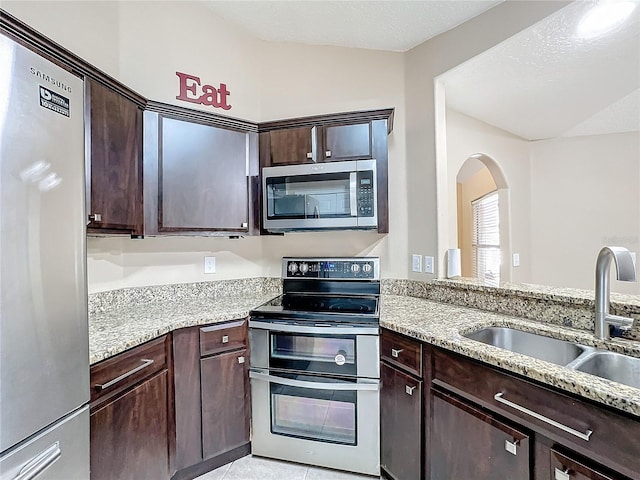 kitchen featuring light stone counters, appliances with stainless steel finishes, vaulted ceiling, dark brown cabinetry, and sink