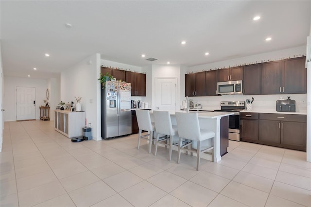 kitchen featuring a breakfast bar, dark brown cabinets, stainless steel appliances, light tile patterned floors, and a center island with sink