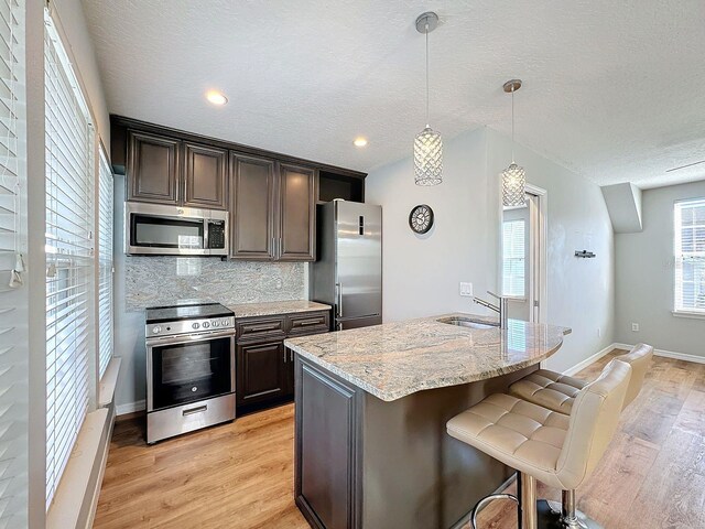 kitchen featuring decorative backsplash, a kitchen island with sink, hanging light fixtures, stainless steel appliances, and light hardwood / wood-style floors