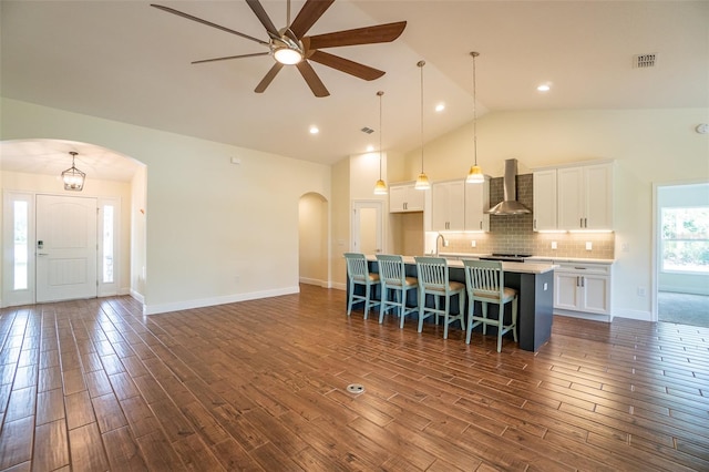 kitchen featuring wall chimney range hood, hanging light fixtures, white cabinetry, a kitchen island with sink, and dark hardwood / wood-style floors