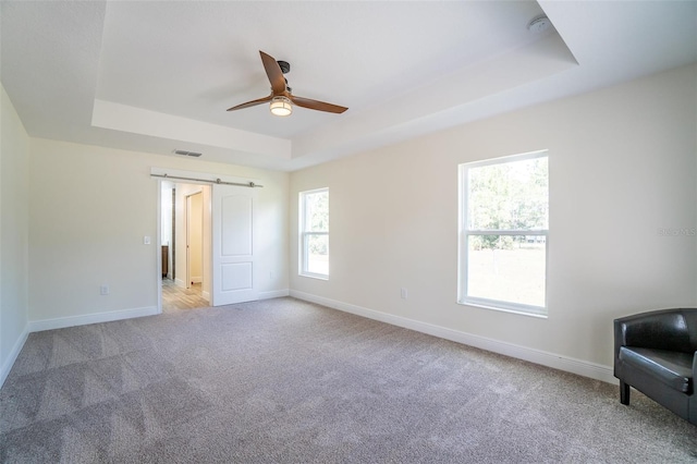 unfurnished bedroom with a barn door, light colored carpet, a tray ceiling, and ceiling fan