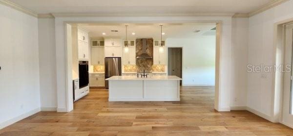 kitchen with wall chimney range hood, stainless steel fridge, white cabinetry, a kitchen island with sink, and pendant lighting