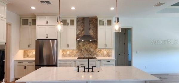 kitchen with stainless steel refrigerator, black electric cooktop, pendant lighting, wall chimney exhaust hood, and light stone counters