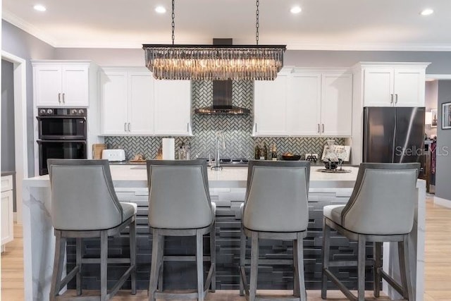 kitchen featuring hanging light fixtures, a center island with sink, white cabinetry, wall chimney exhaust hood, and double oven