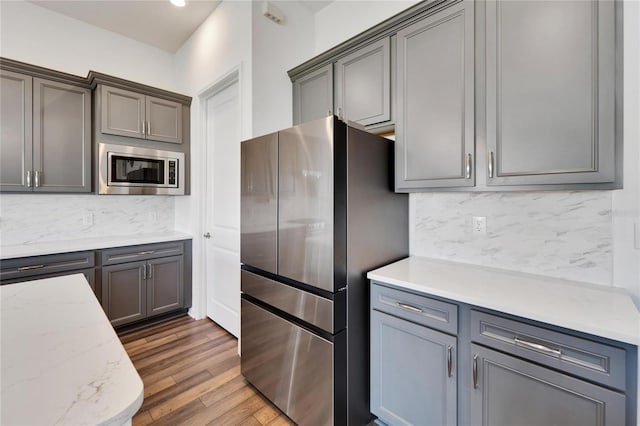 kitchen featuring decorative backsplash, light stone countertops, gray cabinetry, stainless steel appliances, and dark wood-type flooring