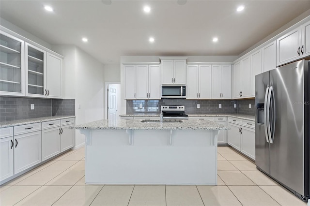 kitchen with light stone countertops, appliances with stainless steel finishes, an island with sink, backsplash, and white cabinets