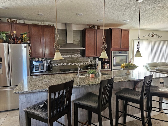 kitchen featuring stone counters, pendant lighting, a kitchen island with sink, appliances with stainless steel finishes, and wall chimney exhaust hood