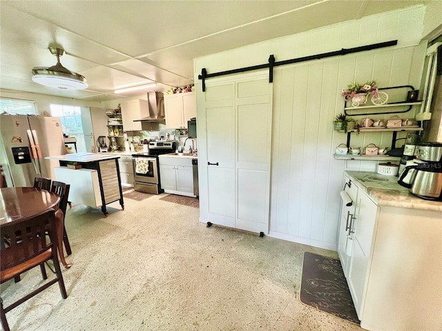 kitchen with wall chimney range hood, white cabinets, appliances with stainless steel finishes, a barn door, and sink