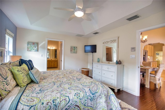 bedroom featuring connected bathroom, dark wood-type flooring, stainless steel refrigerator with ice dispenser, a tray ceiling, and ceiling fan