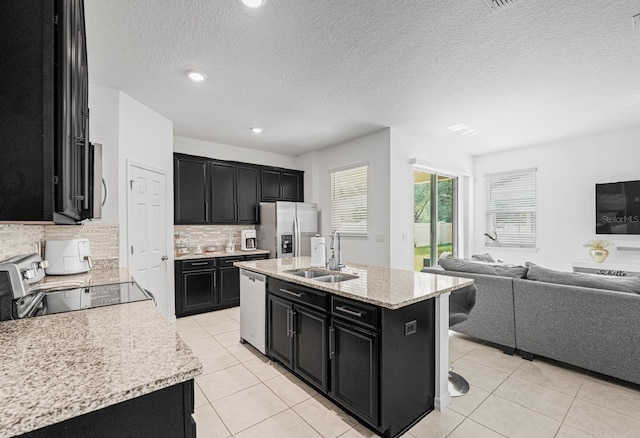 kitchen with sink, a kitchen island with sink, backsplash, light stone countertops, and appliances with stainless steel finishes