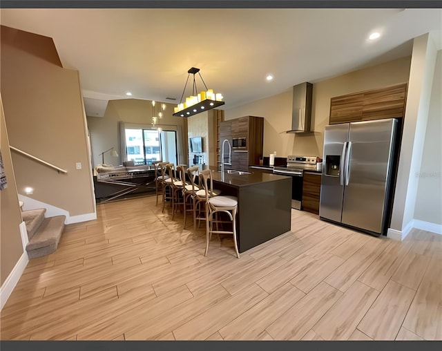 kitchen with hanging light fixtures, stainless steel appliances, wall chimney range hood, a kitchen island with sink, and a breakfast bar