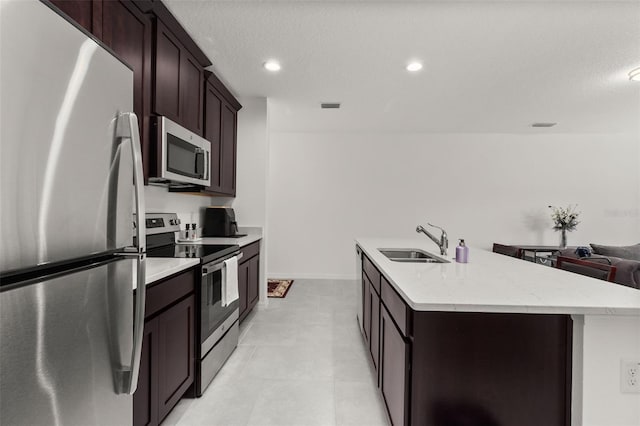 kitchen with stainless steel appliances, a textured ceiling, sink, an island with sink, and dark brown cabinets