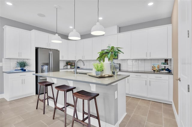 kitchen featuring decorative backsplash, an island with sink, stainless steel fridge with ice dispenser, white cabinetry, and decorative light fixtures