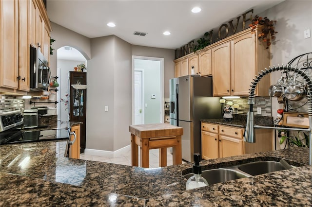 kitchen featuring stainless steel appliances, light brown cabinetry, dark stone countertops, and tasteful backsplash