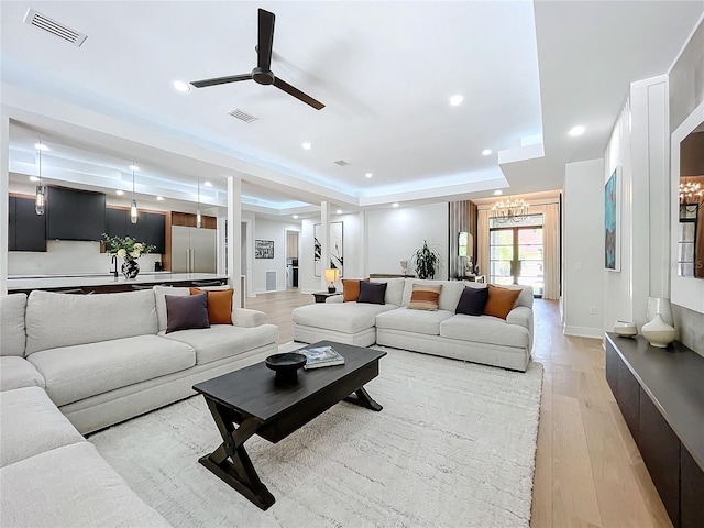 living room with light wood-type flooring, ceiling fan with notable chandelier, and a raised ceiling