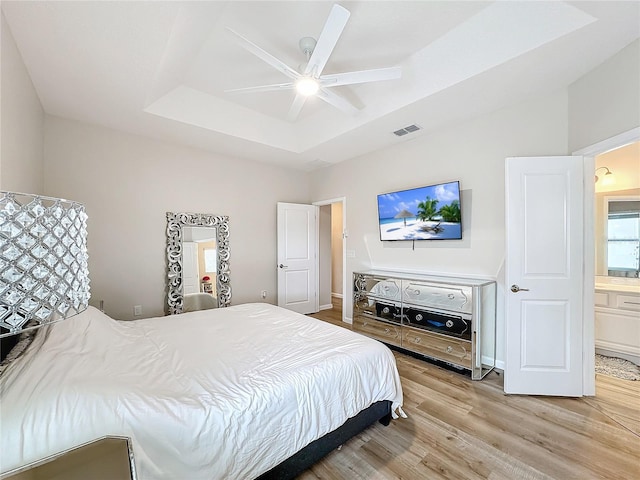 bedroom with light wood-type flooring, a tray ceiling, ensuite bath, and ceiling fan