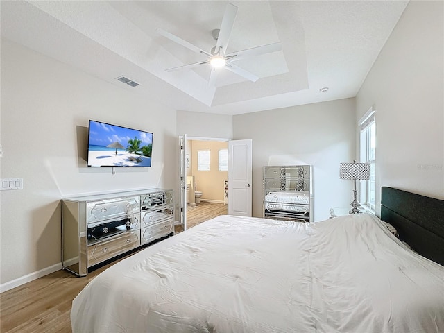 bedroom with a tray ceiling, ensuite bath, ceiling fan, and light wood-type flooring