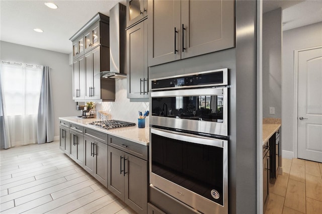 kitchen with light stone counters, appliances with stainless steel finishes, wall chimney exhaust hood, backsplash, and light wood-type flooring