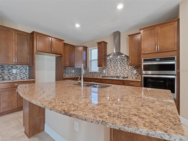 kitchen featuring stainless steel appliances, wall chimney exhaust hood, sink, an island with sink, and light stone countertops