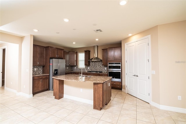 kitchen featuring wall chimney exhaust hood, an island with sink, a kitchen breakfast bar, light stone countertops, and appliances with stainless steel finishes