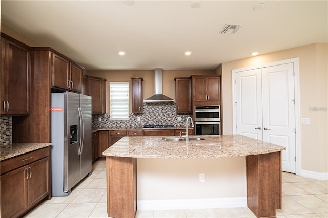 kitchen with stainless steel appliances, sink, an island with sink, wall chimney range hood, and light stone countertops