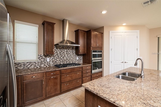 kitchen featuring light stone counters, appliances with stainless steel finishes, dark brown cabinetry, sink, and wall chimney exhaust hood