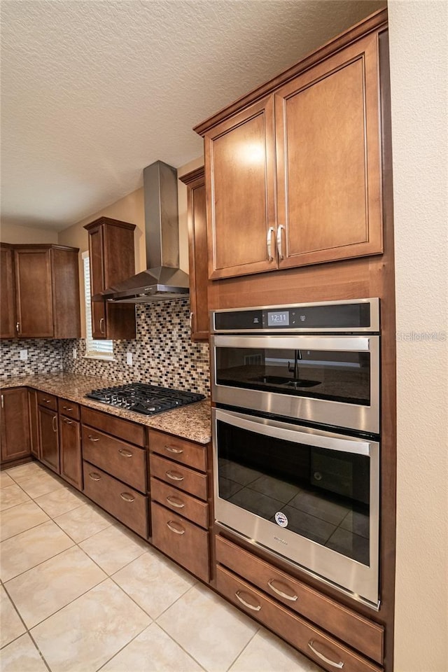 kitchen with light tile patterned flooring, stone countertops, appliances with stainless steel finishes, wall chimney exhaust hood, and backsplash