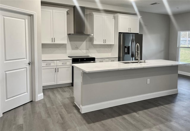 kitchen featuring stainless steel appliances, wall chimney range hood, a center island with sink, and white cabinetry