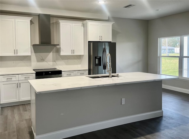 kitchen with wall chimney range hood, white cabinetry, a kitchen island with sink, and stainless steel fridge with ice dispenser