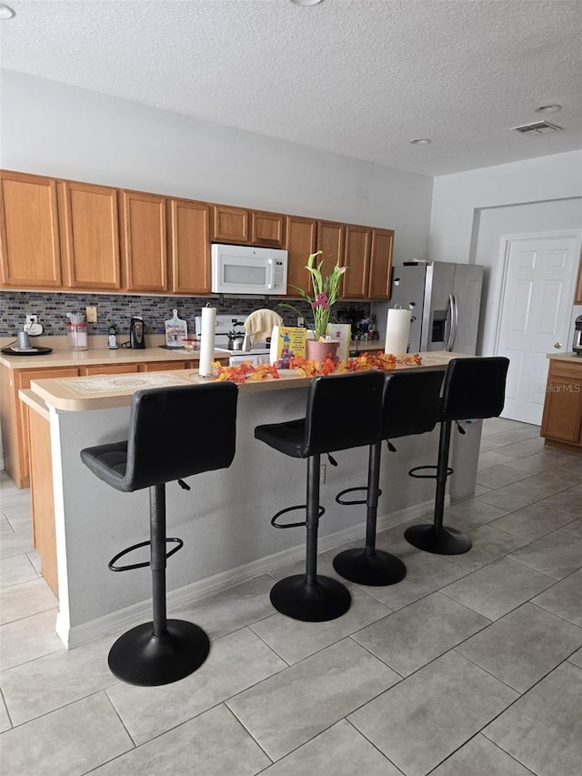 kitchen with stainless steel refrigerator with ice dispenser, tasteful backsplash, a textured ceiling, a center island with sink, and a breakfast bar area