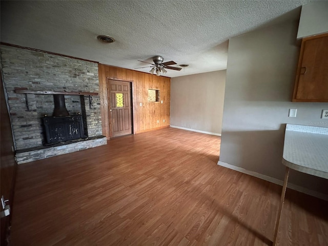 unfurnished living room featuring hardwood / wood-style floors, a wood stove, wooden walls, ceiling fan, and a textured ceiling