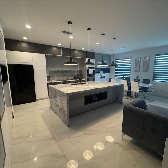 kitchen with white cabinetry, stainless steel double oven, light stone counters, decorative light fixtures, and a kitchen island with sink