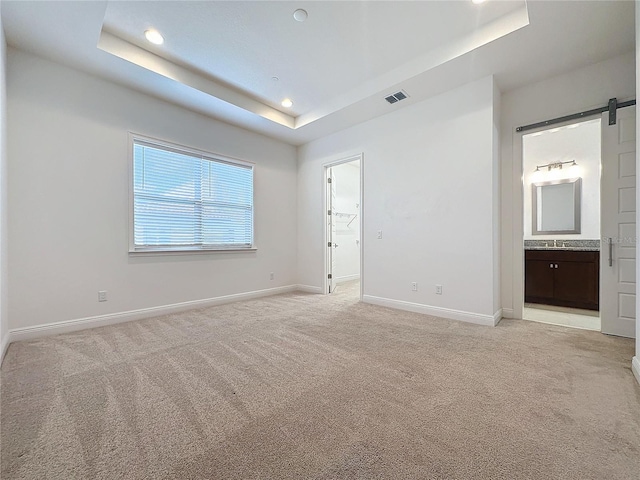 unfurnished bedroom featuring sink, ensuite bath, a barn door, a tray ceiling, and light colored carpet